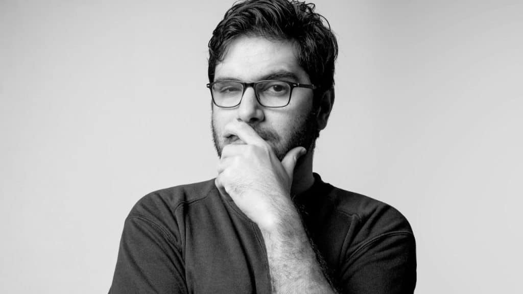 A black and white portrait of a man in glasses, hand on his chin, looking skeptical.