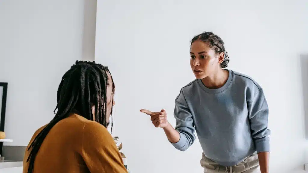 Woman with braided hair points a finger assertively at a man with dreadlocks sitting at a table.