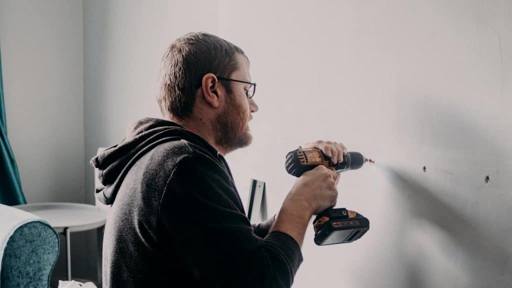 Man in a hoodie and glasses using a cordless drill to make a hole in a white wall in a room.