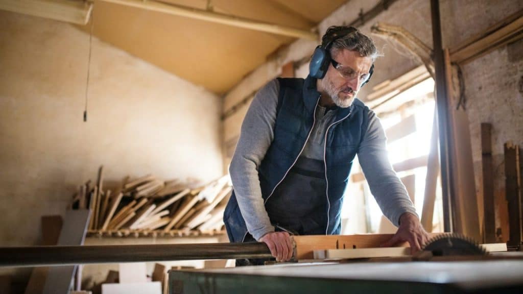 A man wearing safety gear and earmuffs uses a circular saw in a woodworking shop.