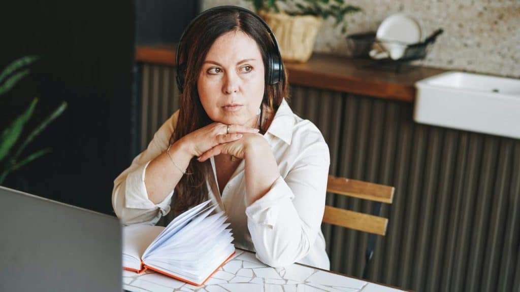 A woman wearing headphones sitting at a table with an open notebook, looking thoughtful.