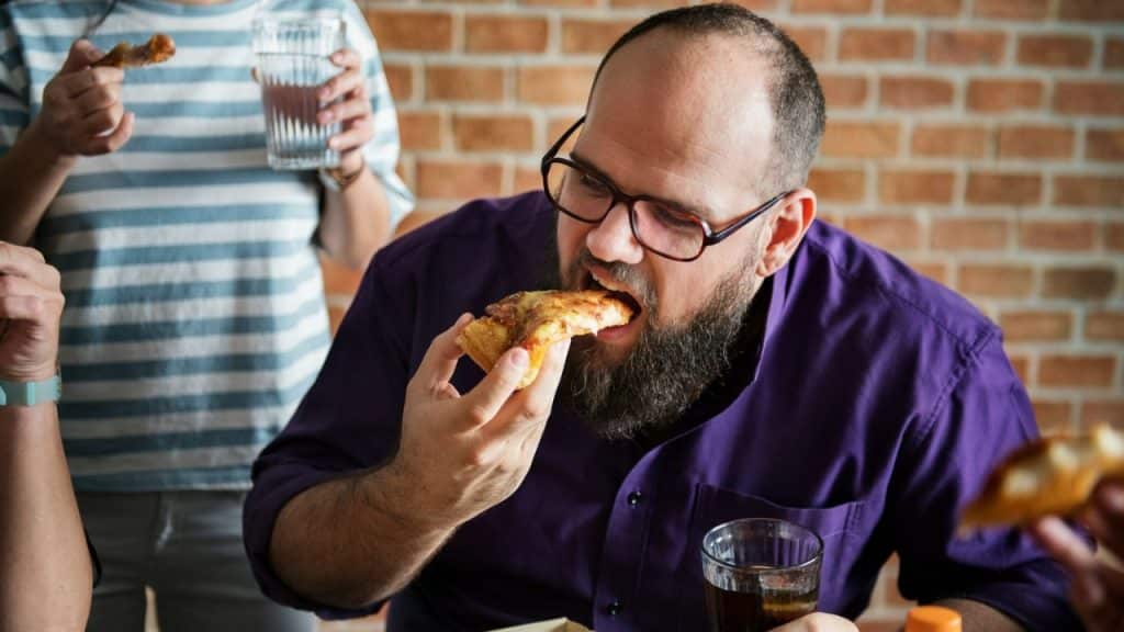 A bearded man with glasses eagerly eats a slice of pizza at a table with other people.