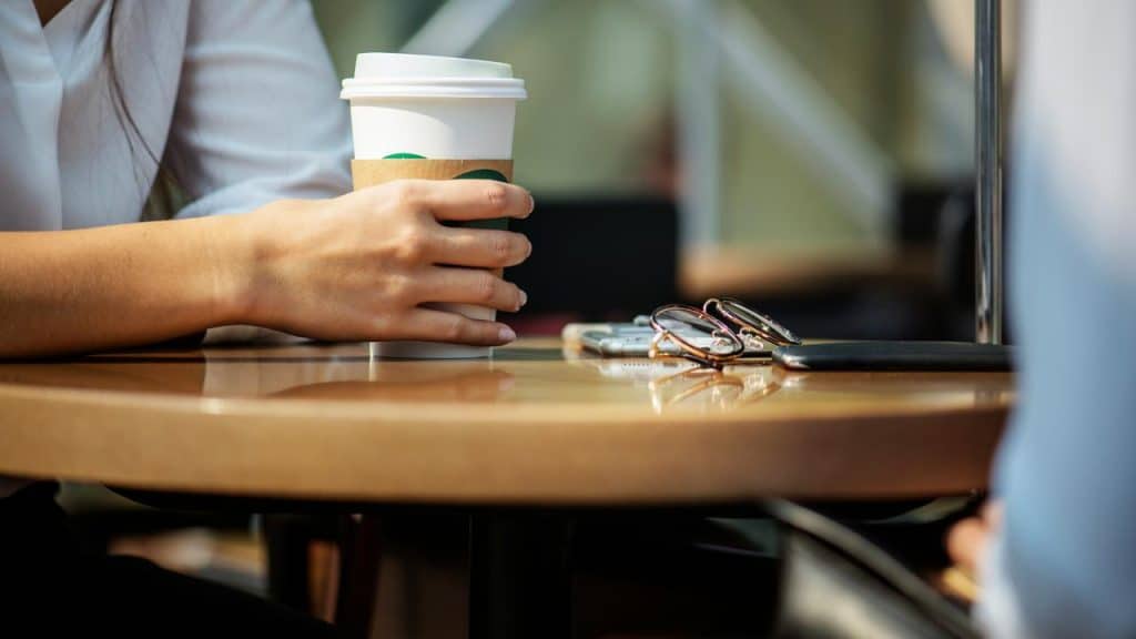 A person holding a coffee cup at a table with glasses and a phone.