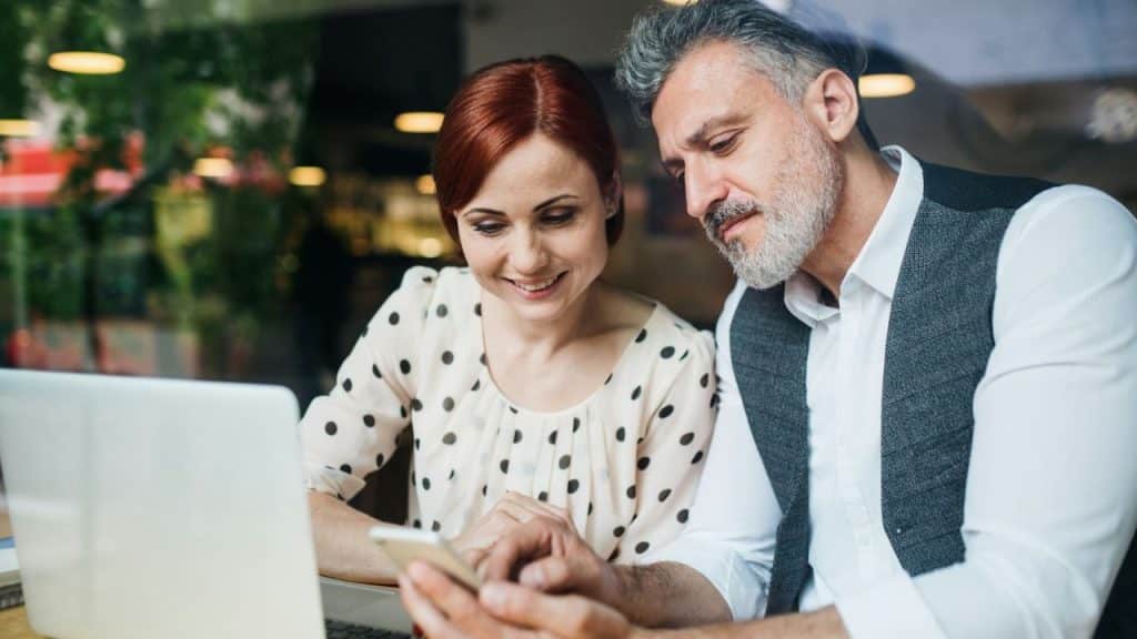 Mature couple looking at a smartphone and laptop together in a cafe.