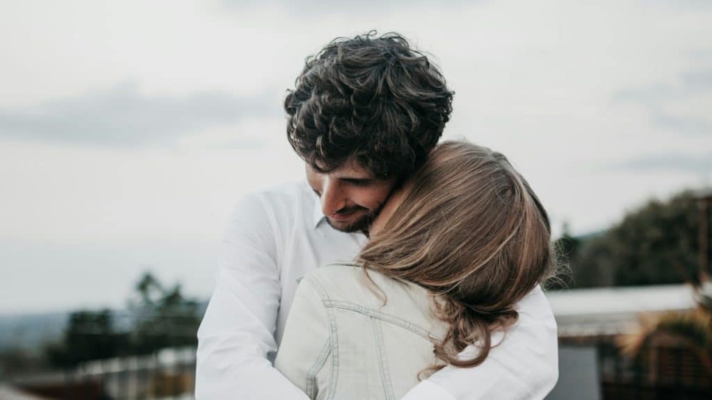 A man with curly hair and a white shirt tightly embraces a woman wearing a light denim jacket.