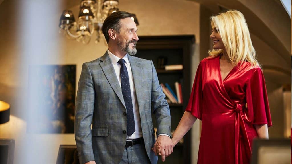 A handsome man and an attractive lady in formal attire holding hands and looking at each other in a well-furnished room.