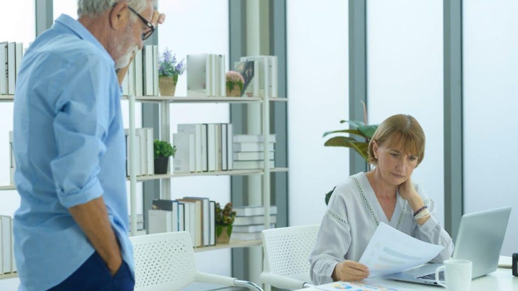An older woman looks over documents while a man stands nearby in an office setting.