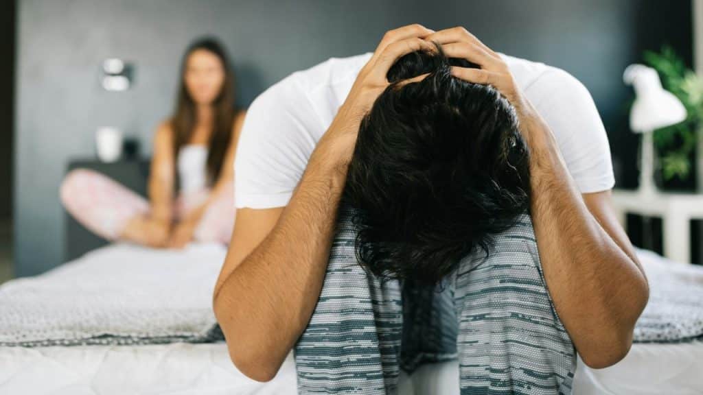 A man sits on the floor with his head in his hands; a woman is out of focus behind him.