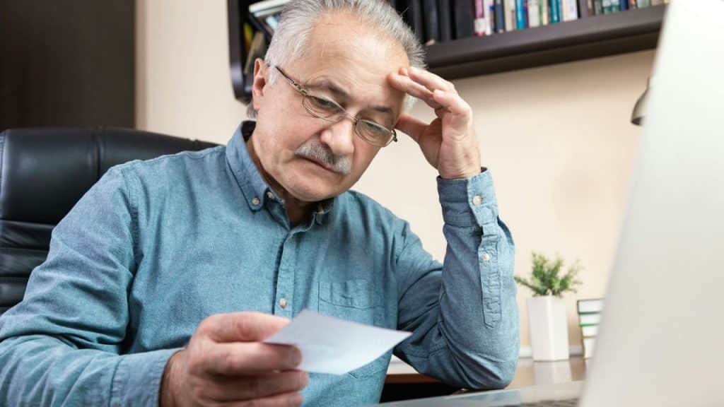 An older man with glasses is sitting at a desk, looking stressed while holding a piece of paper.