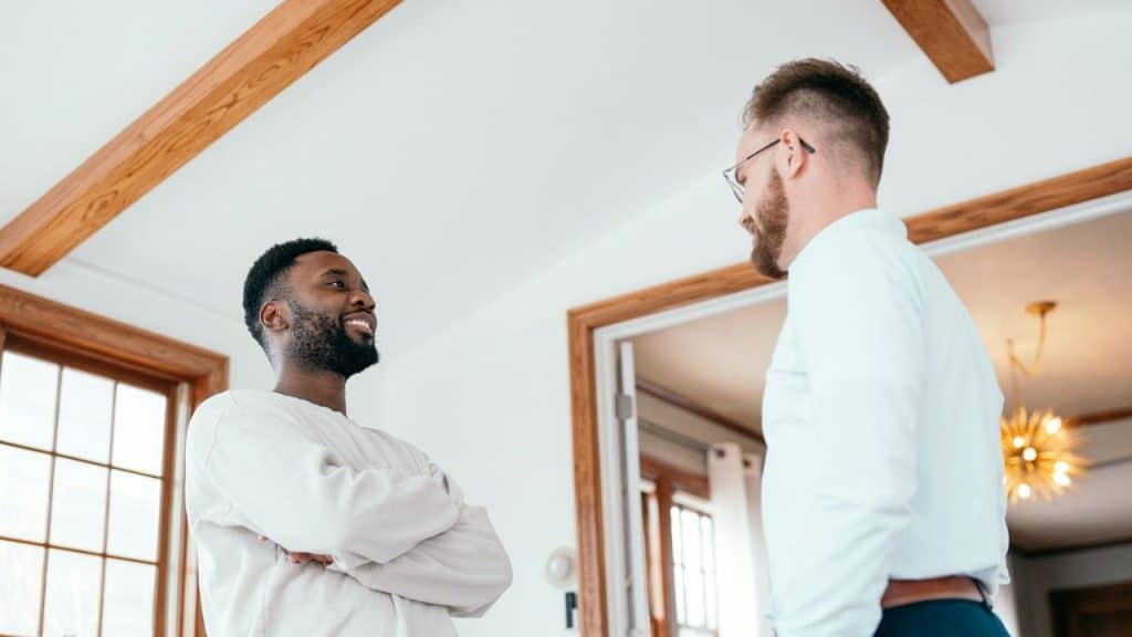 Smiling man with crossed arms talking to another man in a white shirt indoors.