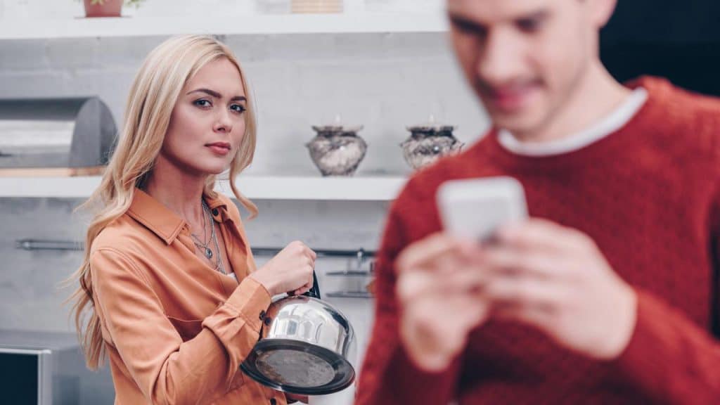 Woman holding a kettle and looking at the camera while a blurred man checks his phone.