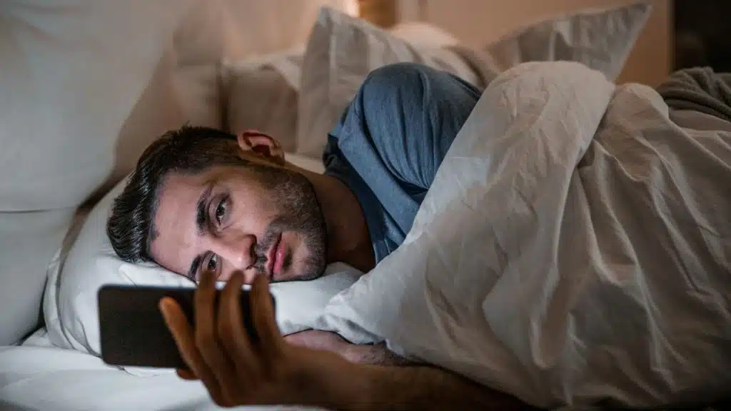 A man lies in bed under covers, looking intently at his phone's screen.