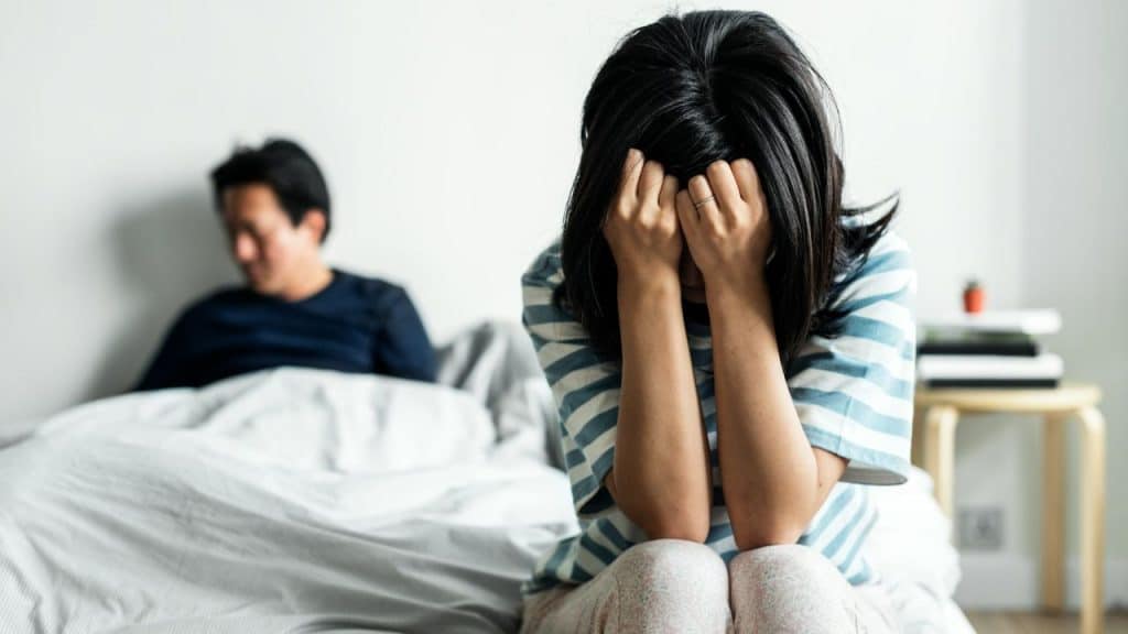 A woman sits on a bed, head in hands, with a man out of focus on the bed behind her.