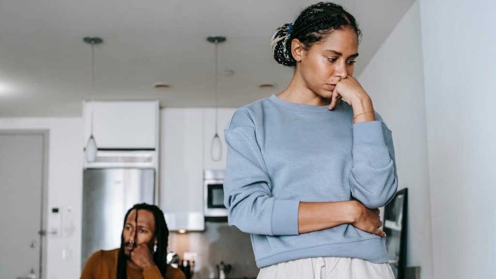 An upset woman stands in the foreground, ignoring a man sitting distressed in the background kitchen.