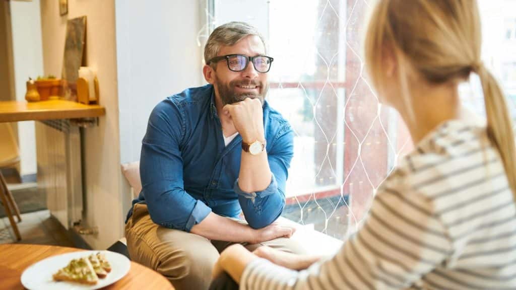 Smiling man in glasses and blue shirt listening to a woman in a cafe.