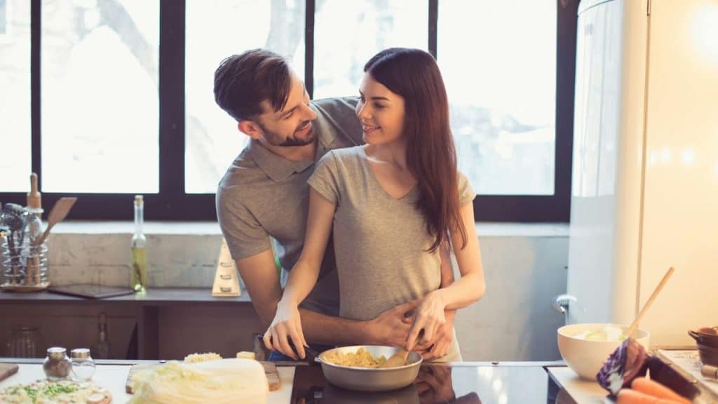 Man and woman embracing and smiling while cooking together in a bright kitchen.