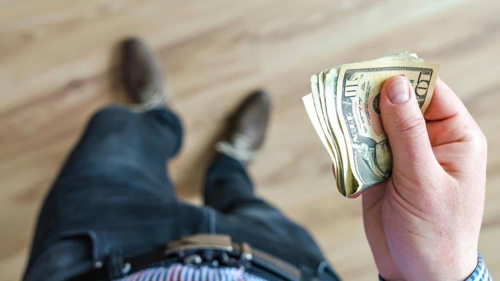 A person holding a small stack of dollar bills while standing on a wooden floor.