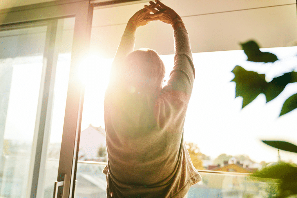 Woman with her back to the camera, standing by a bright window or glass door, stretching her arms up toward the sun.