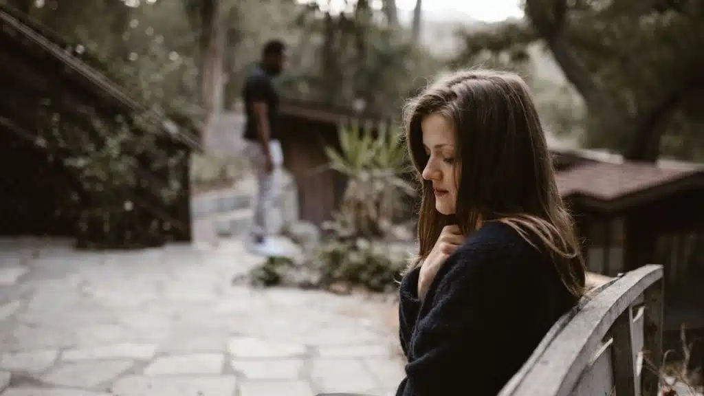 A woman sitting sadly on an outdoor bench with a man standing in the blurry background.