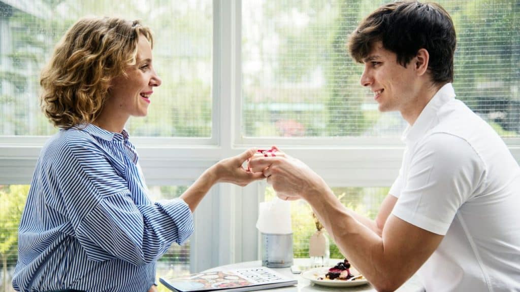 A smiling woman and man sit across a table, gently exchanging a small wrapped gift.