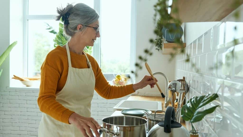 Woman in glasses and apron stirring a pot in a brightly lit kitchen.