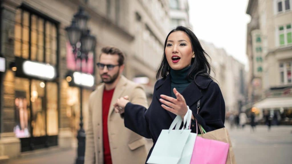 A woman holding shopping bags gestures on a city street.