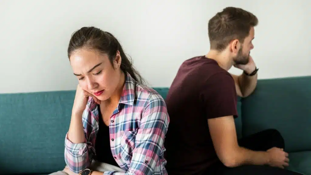 A man and woman sit back-to-back on a teal couch, looking away from each other.