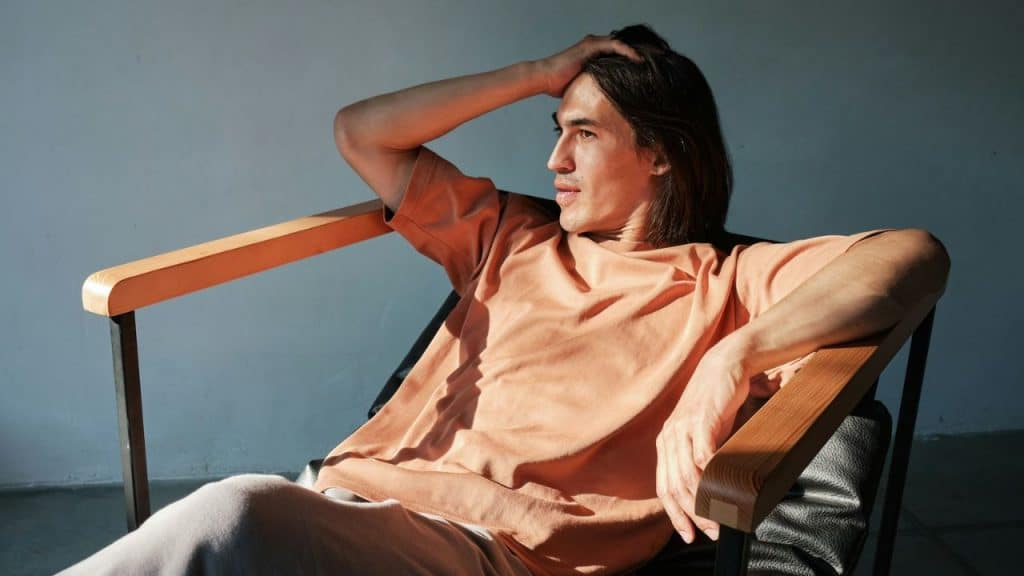 Young man with long hair relaxing in a wooden chair, hand behind his head.