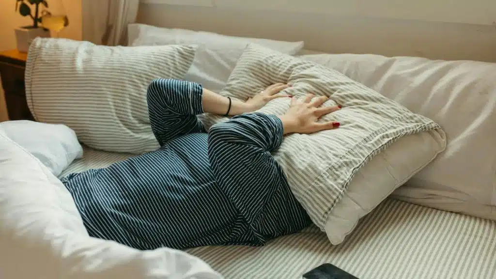 A person lying in bed with a pillow over their face, looking frustrated.