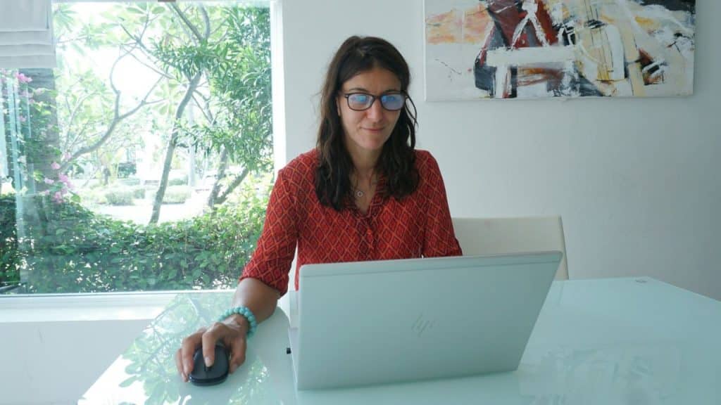 Woman in red patterned shirt and glasses working on a laptop at a glass table.
