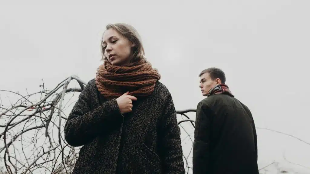 A woman in a thick scarf and coat looks down, while a man in a dark jacket stands behind her.