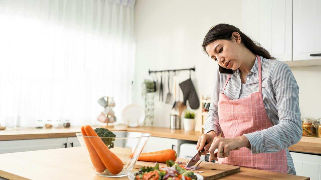 A woman wearing a pink apron chopping vegetables while talking on the phone.