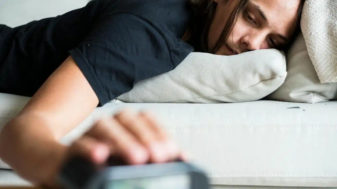 A man lying on a couch reaching for an alarm clock.