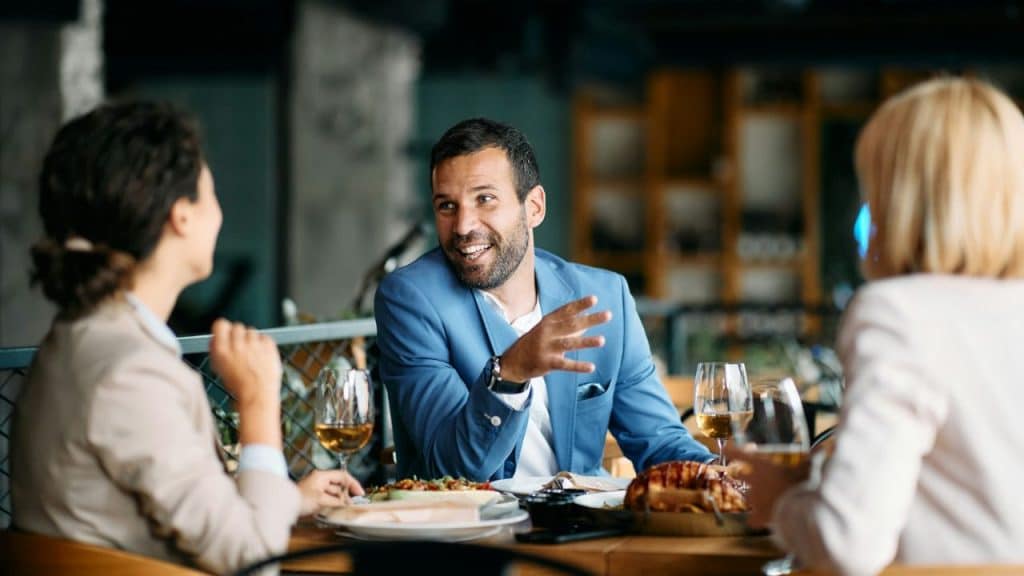 Man in a light blue suit talking to two women over a meal and wine.