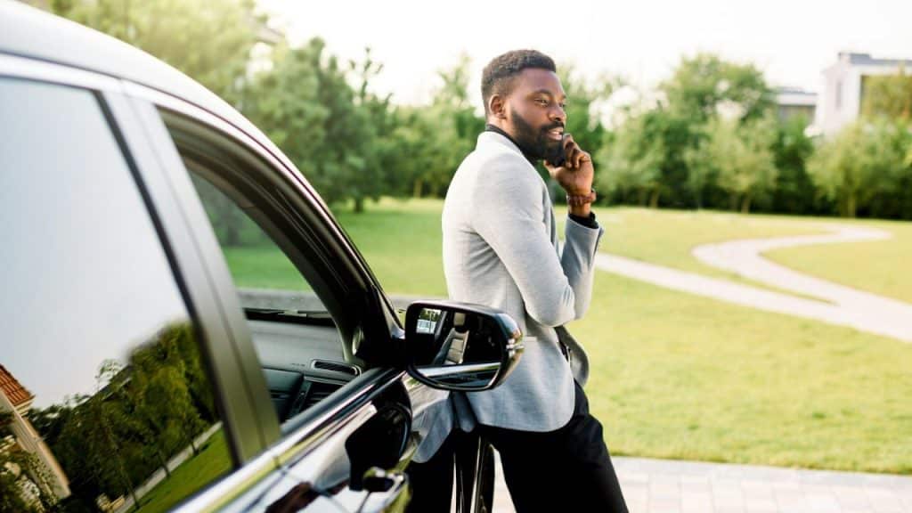 Man in a gray blazer and black pants leans on a black car, talking on a phone.