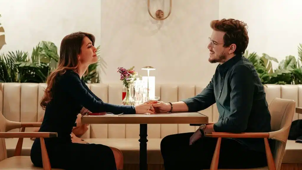 A man and woman sit at a small restaurant table holding hands and smiling.