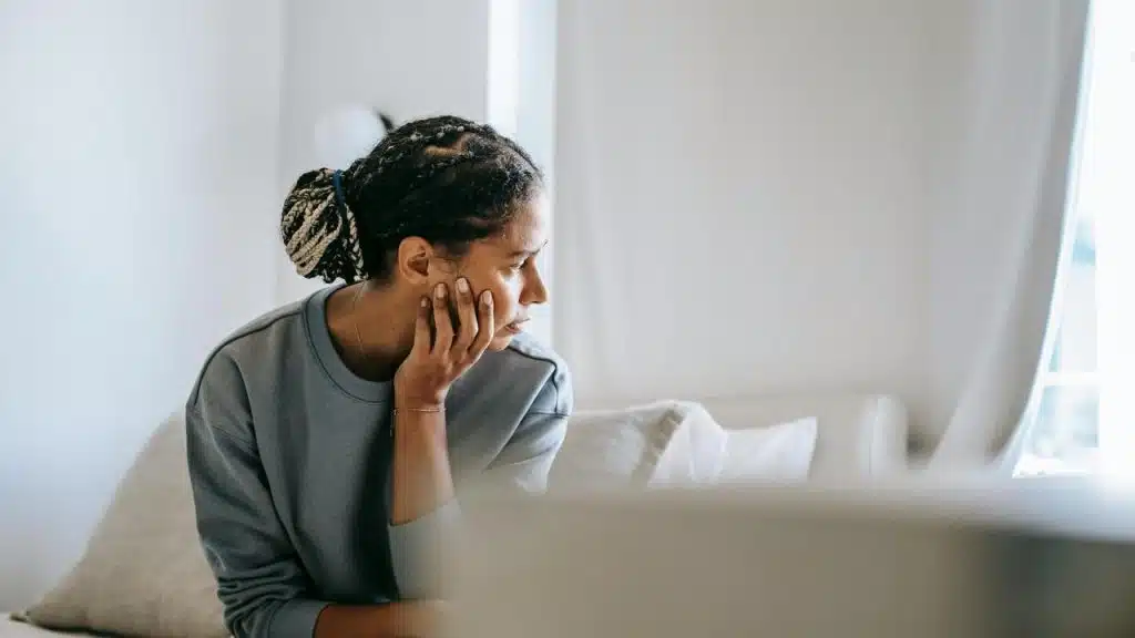 A person sitting on a bed, resting their chin on their hand, appearing pensive.