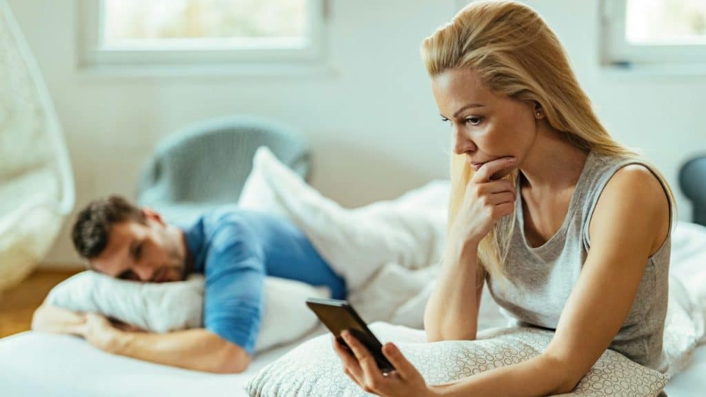 A pensive woman looks at her phone while a man lies in bed in the background.