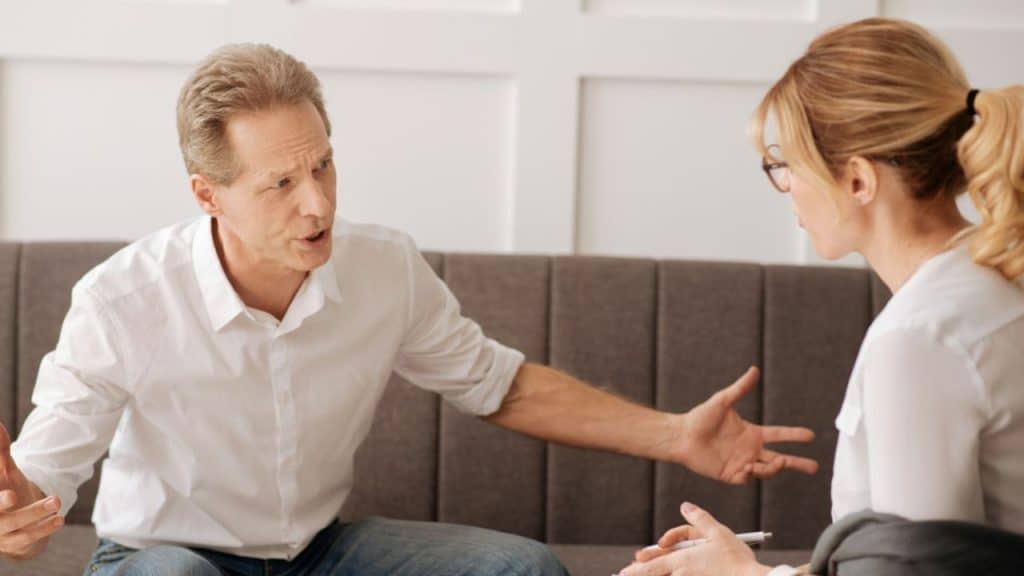 A man in a white shirt speaks animatedly with his hands out toward a seated woman.