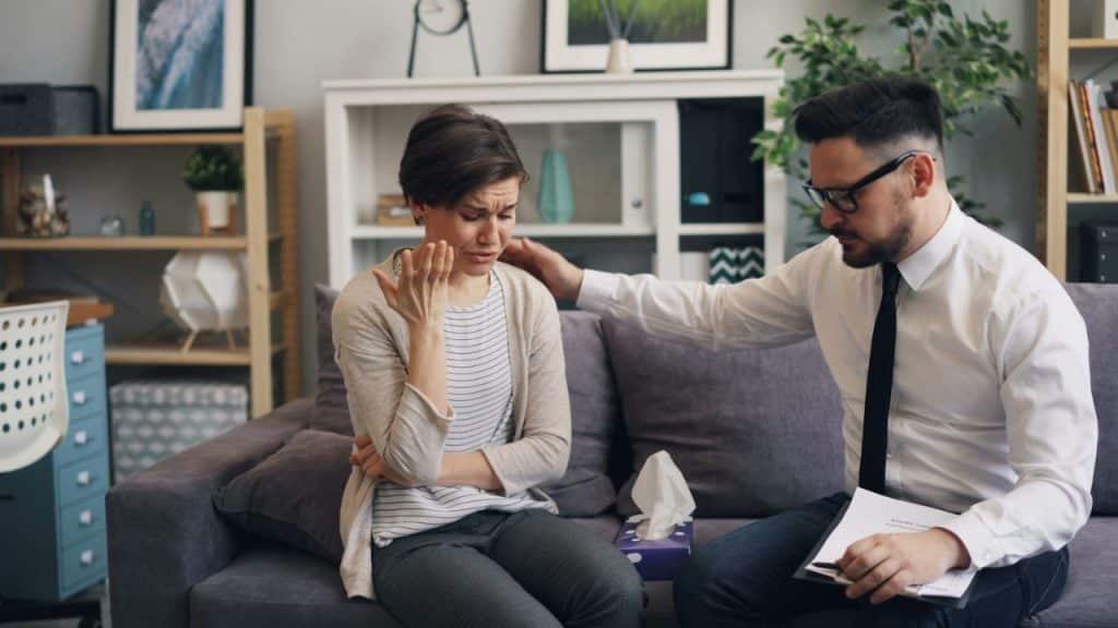 A woman sits on a couch crying while a man in a tie comforts her.