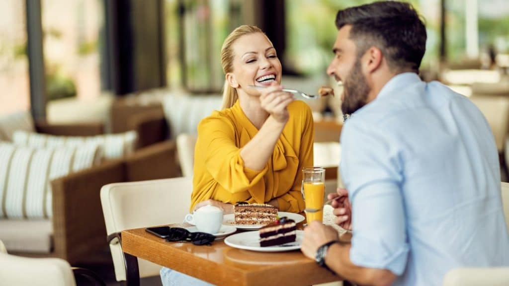 A woman in a yellow shirt feeds a piece of cake to a man at a table.