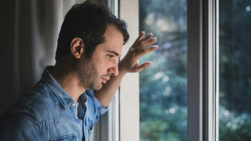 A man in a denim shirt stands by a window, looking outside with one hand on the glass.