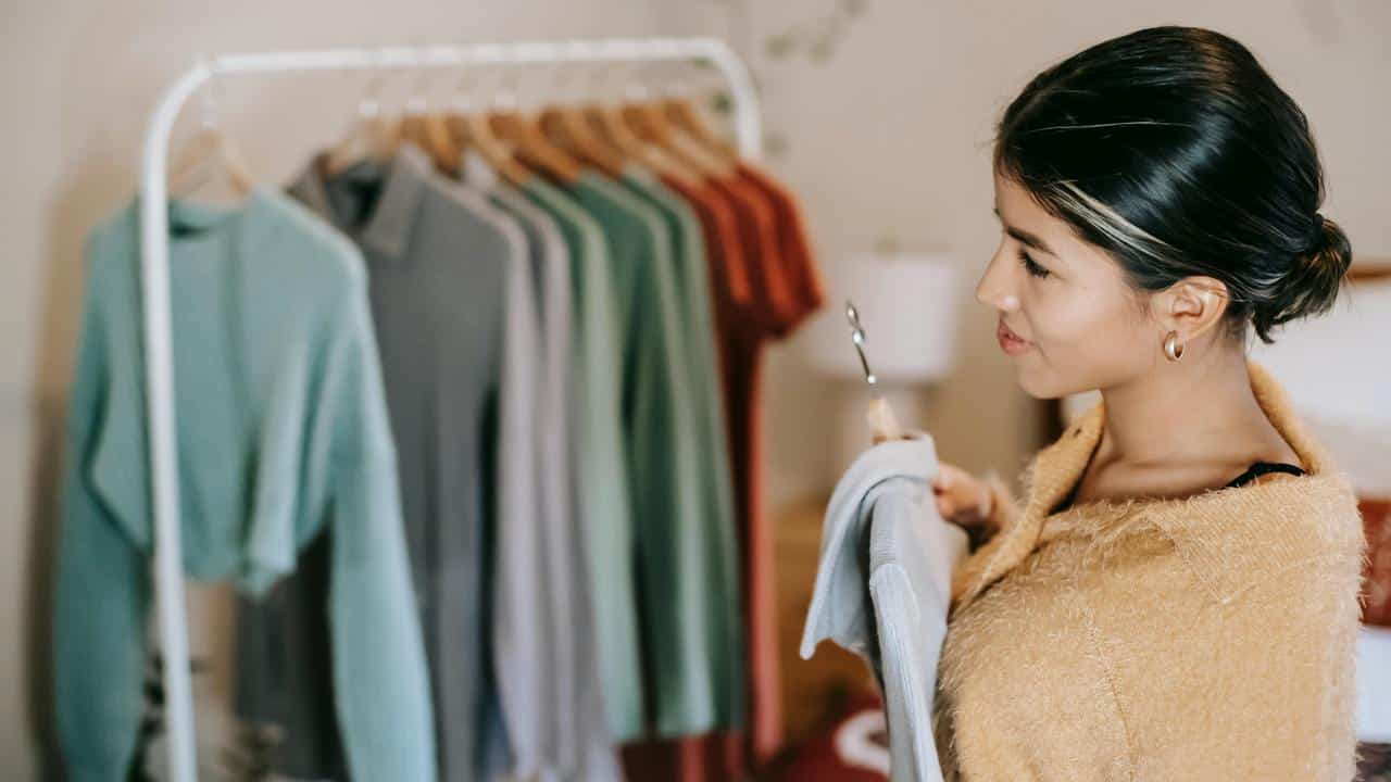 A woman holds a garment on a hanger while standing near a clothing rack.