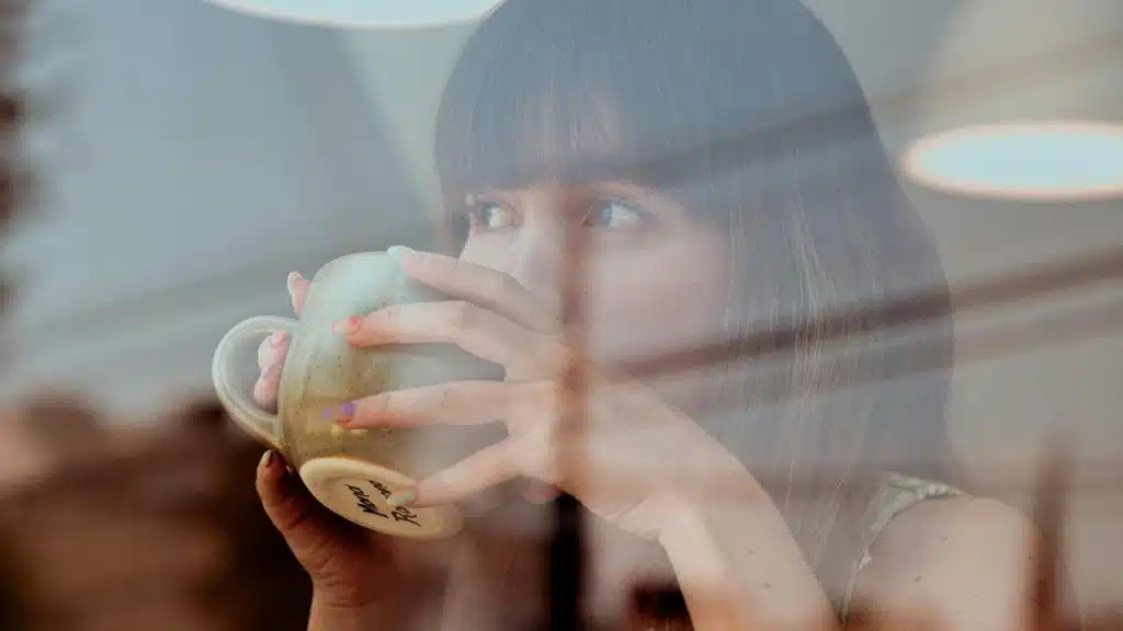 A woman sipping coffee while gazing out through a window with reflections.