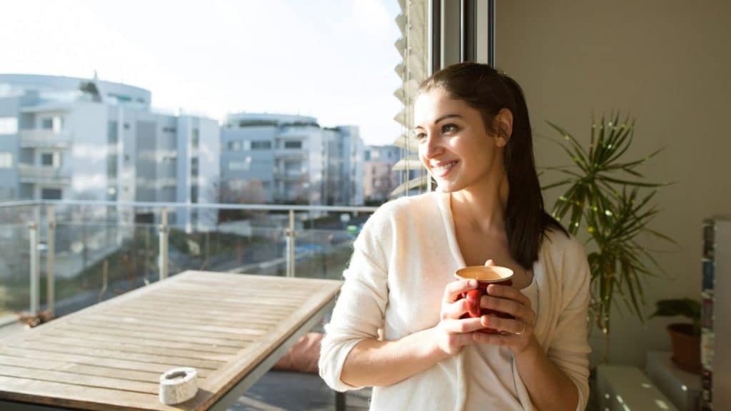 A woman stands by a window holding a red mug and looking out at the city.
