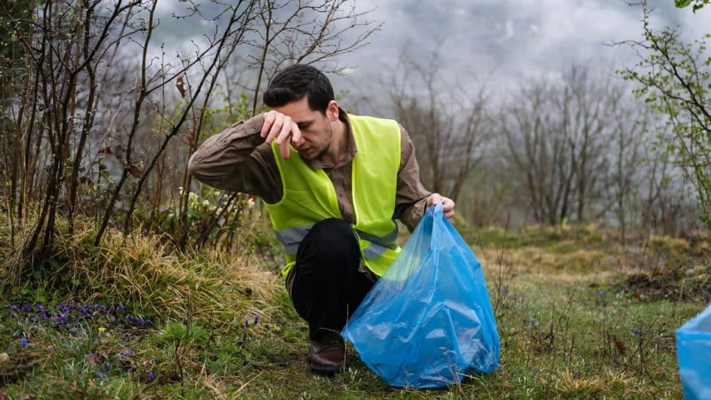 A man in a yellow safety vest kneels outdoors holding a blue plastic bag.