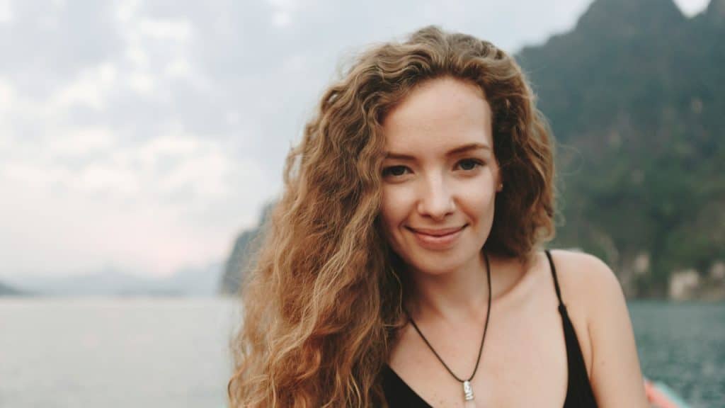 A smiling woman with curly hair standing in front of the sea and looking directly at the camera.