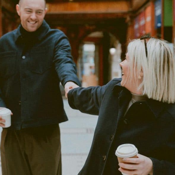 A smiling couple walking indoors holding coffee and reaching for each other.