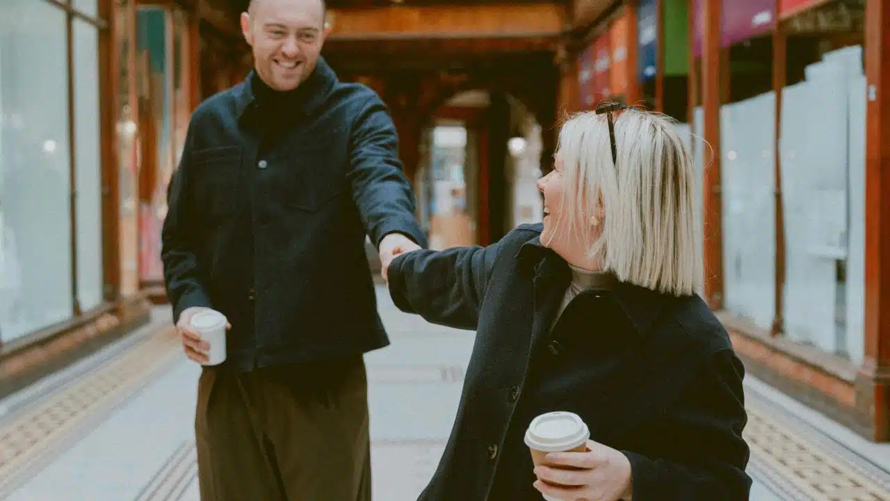A smiling couple walking indoors holding coffee and reaching for each other.