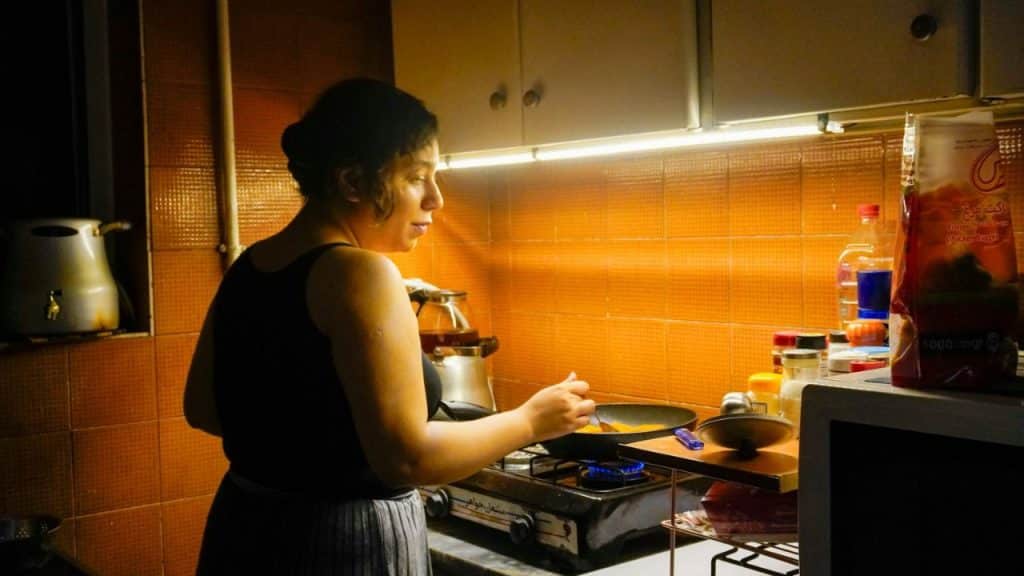 Woman in a tank top cooking on a gas burner in a dimly lit, tiled kitchen.